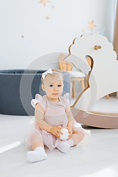 A cute blue-eyed one-year-old baby in a pink dress holds a small white ball in her hands and sits on the floor in the children`s