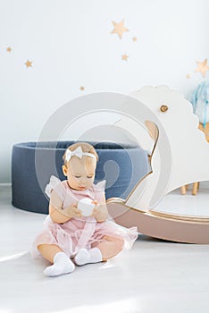 A cute blue-eyed one-year-old baby in a pink dress holds a small white ball in her hands and sits on the floor in the children`s