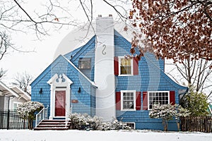 Cute blue painted shingled vintage cottage in the snow with red shutters and white chimney and trim