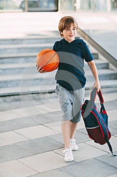 A cute boy with a backpack in one hand and a basketball in the other. The boy in shape in a hurry for a workout but basketball.