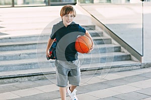 A cute boy with a backpack in one hand and a basketball in the other. The boy in shape in a hurry for a workout but basketball.