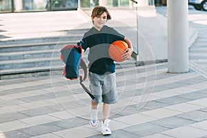 A cute boy with a backpack in one hand and a basketball in the other. The boy in shape in a hurry for a workout but basketball.