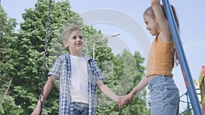 Cute boy and beautiful girl with long hair holding hands and talking near the swing, smiling happily. A couple of happy