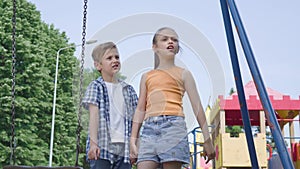 Cute boy and beautiful girl with long hair pointing away holding hands near the swing, smiling happily outdoors. A