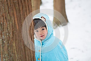 Cute boy in blue cloak playing hide and seek in forest after snow