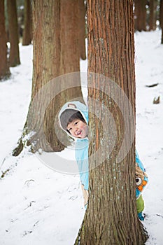 Cute boy in blue cloak playing hide and seek in forest after snow
