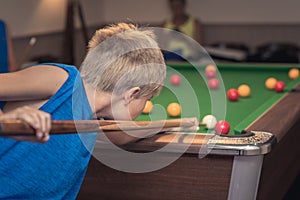 Cute boy in blue t shirt plays billiard or pool in club. Young Kid learns to play snooker. Boy with billiard cue strikes the ball
