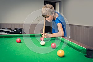 Cute boy in blue t shirt plays billiard or pool in club. Young Kid learns to play snooker. Boy with billiard cue strikes the ball