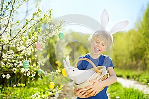 Cute boy in bunny ears holding wicker basket with white toy rabbit during hunt for colorful eggs in spring park on Easter day.