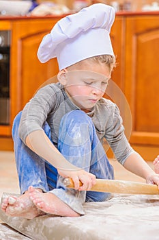 Cute boy in chef`s hats sitting on the kitchen floor soiled with flour, playing with food, making mess and having fun photo