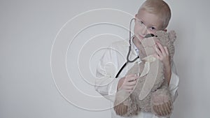 Cute boy in a doctor`s lab coat listening to a teddy bear with a stethoscope