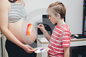 Cute boy drawing rainbow on pregnant belly his mother. Baby birth expecting time and belly painting