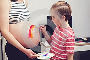 Cute boy drawing rainbow on pregnant belly his mother. Baby birth expecting time and belly painting