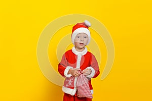 A cute boy dressed like Santa Claus looks at the camera in surprise, in his hands a bag with gifts.