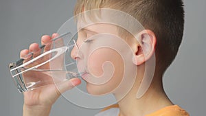 Cute boy drinks clean water from a glass on a gray background. portrait of a child with a glas