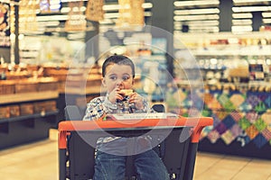 Cute boy eating a bun or pita bread in a supermarket in the cart. Hungry child in the store. toned