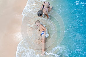 Cute boy and girl having fun on the sunny tropical beach. Lying on sand, wonderful waves around them. View from above.
