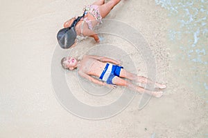 Cute boy and girl having fun on the sunny tropical beach. Lying on sand, wonderful waves around them. View from above.