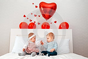 Cute boy and girl sitting together on a white bed and holding hands. Red balloons hang over the bed. The Concept Of Valentine`s