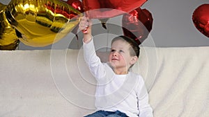 Cute boy having fun, indulging or playing on a light sofa with heart-shaped balloons. Valentine`s Day. Slow motions