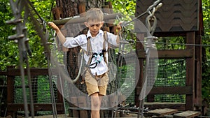 Cute boy having fun while walking on wobbly rope bridge at adventure park. Active childhood, healthy lifestyle, kids playing