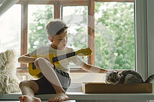 Cute boy learns to play the Ukulele guitar on the windowsill near the window