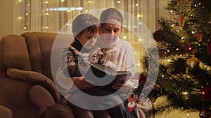 Cute boy with mother sitting armchair next to Christmas tree and reading big book. Families and children celebrating