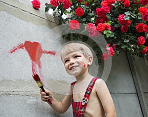 Cute boy in overalls draws a heart on the fence with red paint