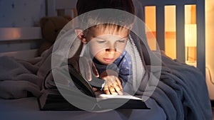 Cute boy in pajamas reading a book with flashlight in bed. Children education, development, secrecy, reading books