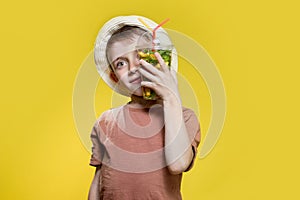 Cute boy in panama drinking mojito cocktail from plastic cup over yellow studio background