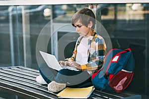 A cute boy in a plaid shirt is sitting on a bench with a laptop and typing on the keyboard, next to a backpack. The student is