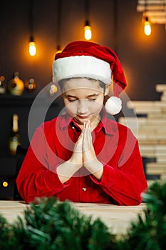 Cute boy praying and dreaming of a gift for Christmas in Santa red hat