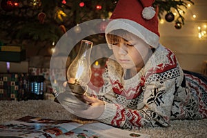 A cute boy in a red hat is reading a children`s book near a Christmas tree and holding a vintage lamp