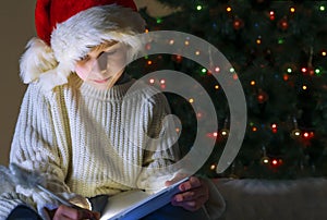 Cute boy in red santa hat and white sweater writing letter to Santa on the background of a Christmas tree decorated with lights