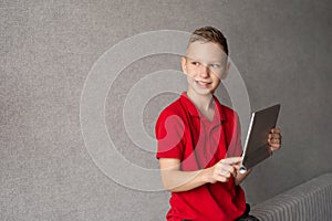 A cute boy in a red T-shirt holds a tablet in his hands and looks away