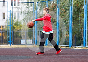 Cute boy in red t shirt plays basketball on city playground. Active teen enjoying outdoor game with orange ball. Hobby