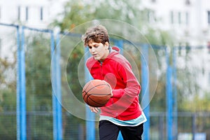 Cute boy in red t shirt plays basketball on city playground. Active teen enjoying outdoor game with orange ball. Hobby,