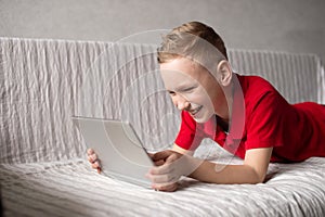 A cute boy in a red T-shirt with a tablet in his hands is lying on the bed. Play on a tablet