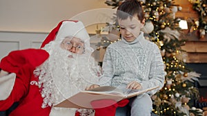 Cute boy and Santa talking and reading book on New Year`s day near decorated tree