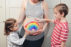 Cute boy with sister drawing rainbow on pregnant belly their mother. Baby birth expecting time and belly painting