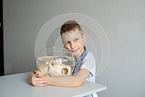 A cute boy sits near a transparent aquarium in the room and hugs an aquarium with fish