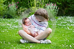 Cute boy sitting in a beautiful summer garden full with flowers holding his newborn sister