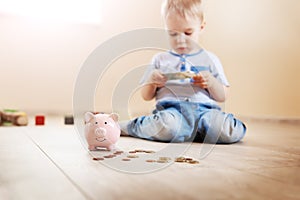 Cute boy sitting on the floor indoor and inserting euro coins in piggybank.