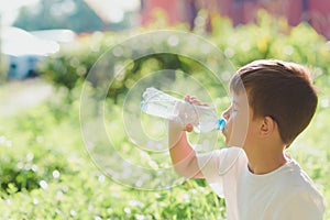 Cute boy sitting on the grass drinks water from a bottle in the summer at sunset. Child quenches thirst on a hot day