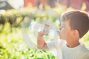 Cute boy sitting on the grass drinks water from a bottle in the summer at sunset. Child quenches thirst on a hot day