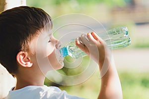 Cute boy sitting on the grass drinks water from a bottle in the summer at sunset. Child quenches thirst on a hot day