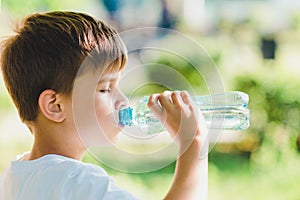 Cute boy sitting on the grass drinks water from a bottle in the summer at sunset. Child quenches thirst on a hot day