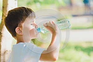 Cute boy sitting on the grass drinks water from a bottle in the summer at sunset. Child quenches thirst on a hot day