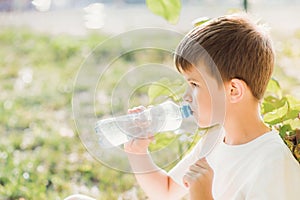 Cute boy sitting on the grass drinks water from a bottle in the summer at sunset. Child quenches thirst on a hot day