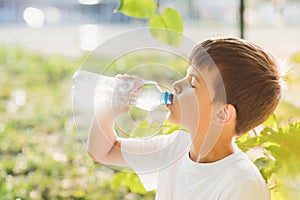 Cute boy sitting on the grass drinks water from a bottle in the summer at sunset. Child quenches thirst on a hot day